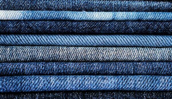 A close-up shot of a stack of Denim | Denim Fabric | Denim Fabric UK displaying various shades and textures, neatly folded and layered on top of each other. The denim fabric ranges in color from light blue to dark indigo, showcasing different patterns and weaves.