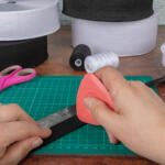 A person holding a piece of elastic along a ruler on a cutting mat, preparing to cut it with a rotary cutter. Scissors, spools of white and black elastic, and a spool of thread are on the table.