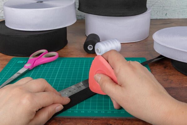A person holding a piece of elastic along a ruler on a cutting mat, preparing to cut it with a rotary cutter. Scissors, spools of white and black elastic, and a spool of thread are on the table.