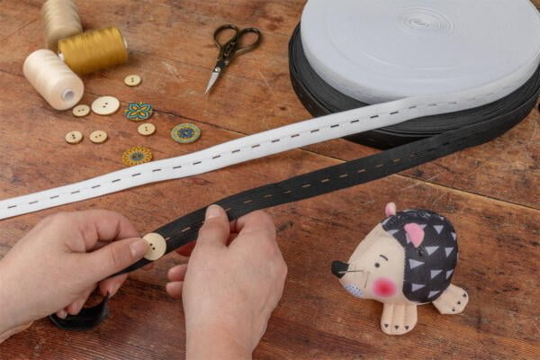 A person is attaching a button to a black elastic band at a wooden table. Nearby are threads, fabric-covered buttons, embroidery, scissors, and white elastic. A small, hedgehog-shaped pincushion sits on the table.