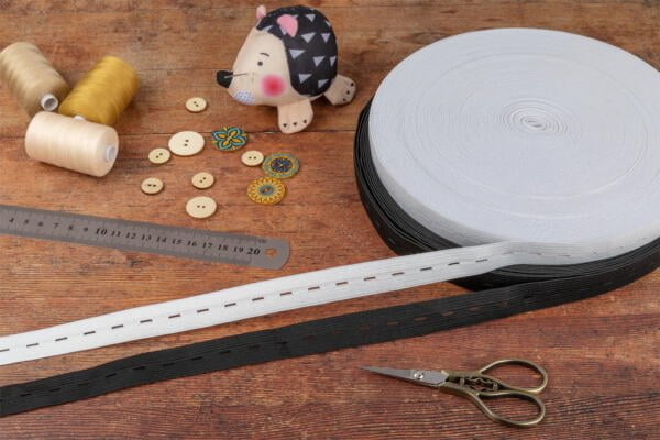 A table with sewing supplies including spools of thread, a ruler, buttons, scissors, rolls of elastic bands, and a hedgehog pin cushion. The wooden surface provides a rustic backdrop.