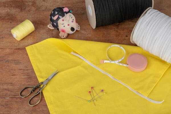 A sewing setup displays a piece of yellow fabric, a pair of scissors, assorted pins, a measuring tape, and yellow and white spools of thread on a wooden surface. A pincushion shaped like a hedgehog rests nearby, alongside a pink retractable measuring tape.