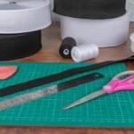 A crafting workspace with green cutting mat, pair of pink-handled scissors, metal ruler, piece of black elastic, pink tailor's chalk, and various rolls of black and white elastic bands and threads in the background.