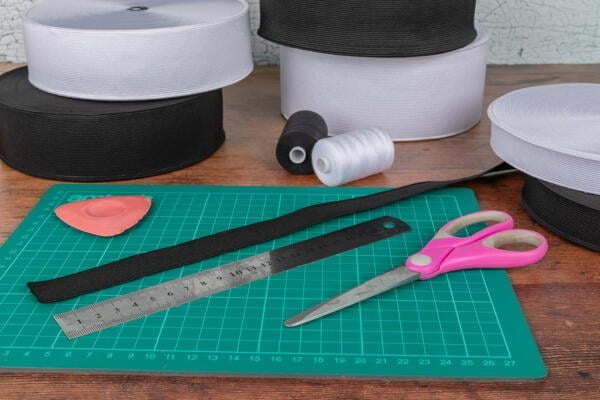 A crafting workspace with green cutting mat, pair of pink-handled scissors, metal ruler, piece of black elastic, pink tailor's chalk, and various rolls of black and white elastic bands and threads in the background.