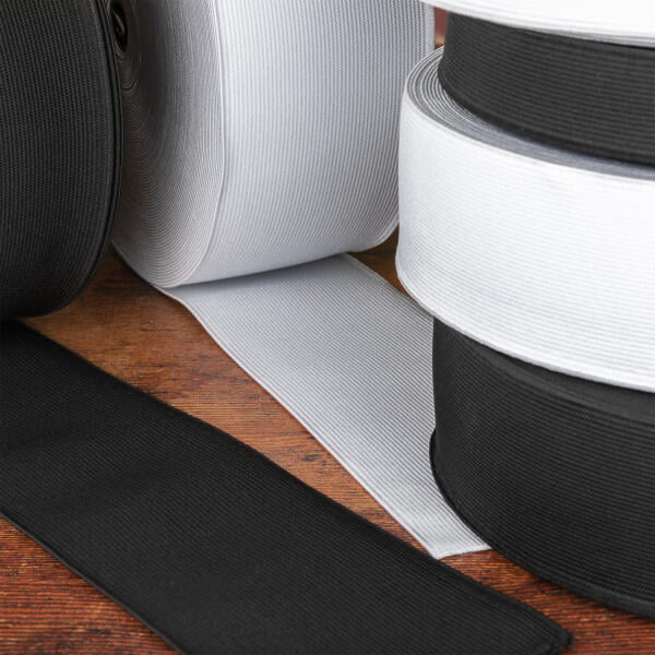 Rolls of elastic fabric in black and white are displayed on a wooden surface. The rolls are unspooling slightly, showing the versatility and texture of the elastic material. The background is rustic, contrasting with the smooth and stretchy fabric.