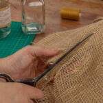 A person is cutting a piece of burlap fabric with scissors on a wooden table. Nearby, a mason jar wrapped in lace, another uncapped glass bottle, a spool of brown thread, and a green cutting mat are visible. The scene suggests a crafting or DIY project.