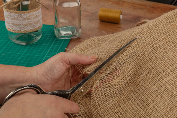 A person is cutting a piece of burlap fabric with scissors on a wooden table. Nearby, a mason jar wrapped in lace, another uncapped glass bottle, a spool of brown thread, and a green cutting mat are visible. The scene suggests a crafting or DIY project.