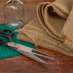 A pair of large scissors, a cutting mat, two glass jars decorated with lace, and a roll of burlap fabric are placed on a wooden table, suggesting a craft or sewing project.