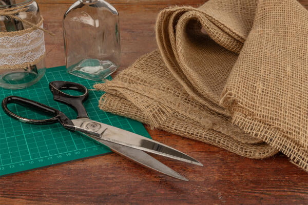 A pair of large scissors, a cutting mat, two glass jars decorated with lace, and a roll of burlap fabric are placed on a wooden table, suggesting a craft or sewing project.