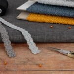 A crafting scene with various sewing materials: stacked fabrics in gray, yellow, and blue, spools of black and white thread, lace trims in gray, pins with orange heads, scissors with ornate handles, and a blue bobbin on a wooden surface.