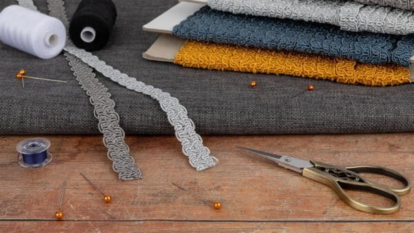 A crafting scene with various sewing materials: stacked fabrics in gray, yellow, and blue, spools of black and white thread, lace trims in gray, pins with orange heads, scissors with ornate handles, and a blue bobbin on a wooden surface.