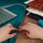 A close-up of hands working with teal fabric on a wooden table. Nearby are spools of colored ropes in teal, red, white, and navy, wrapped around cardboard. Spools of teal thread and a few pink pins are also visible.