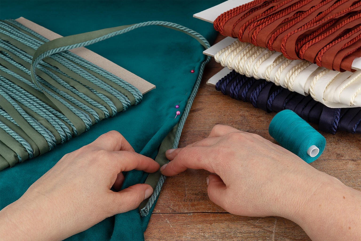 A close-up of hands working with teal fabric on a wooden table. Nearby are spools of colored ropes in teal, red, white, and navy, wrapped around cardboard. Spools of teal thread and a few pink pins are also visible.