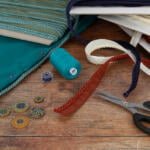 A crafting scene featuring various materials: teal and off-white fabric, spools of thread, a pair of scissors, decorative buttons, and trim with red and cream edges. The items are arranged on a wooden surface, suggesting a project in progress.