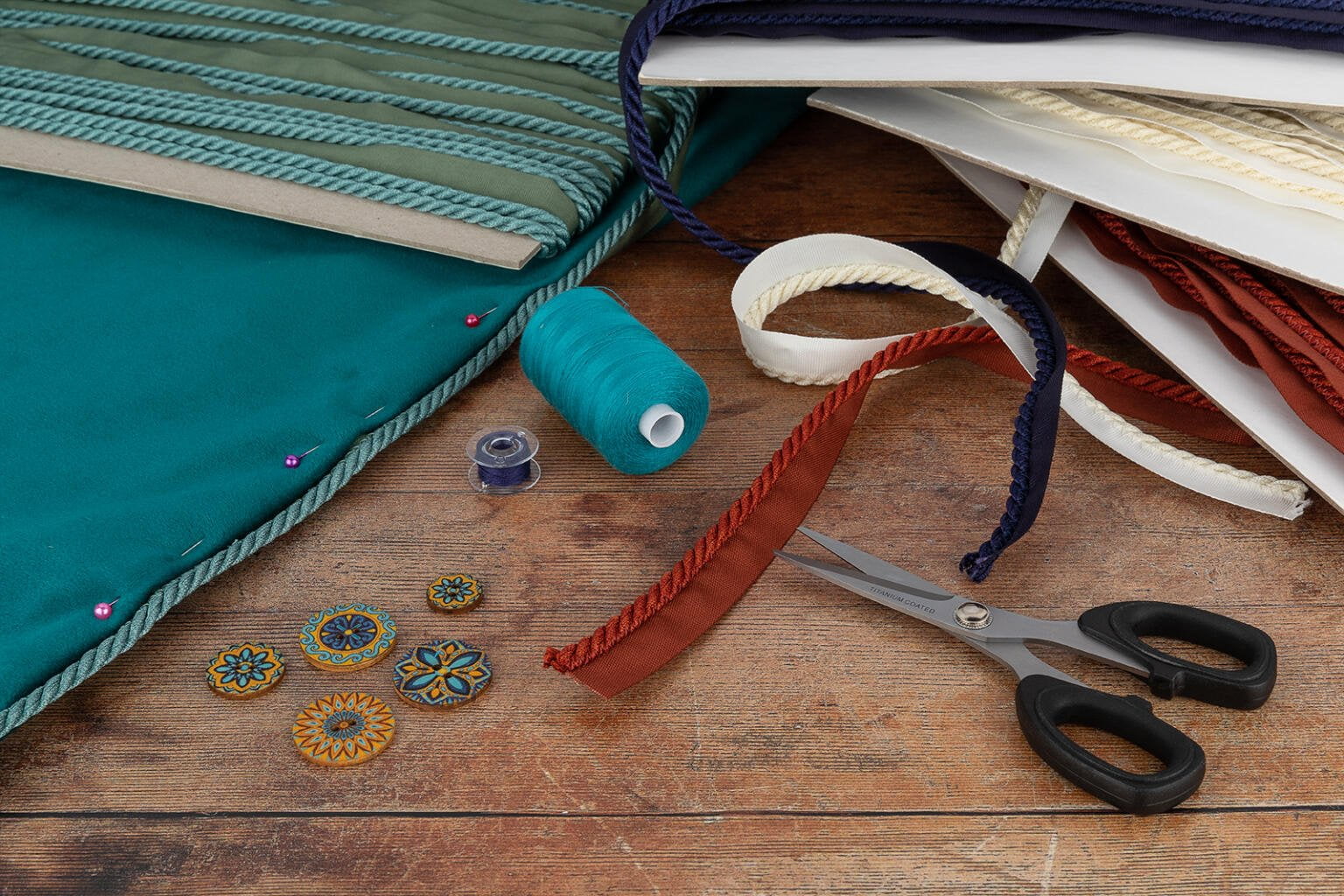 A crafting scene featuring various materials: teal and off-white fabric, spools of thread, a pair of scissors, decorative buttons, and trim with red and cream edges. The items are arranged on a wooden surface, suggesting a project in progress.