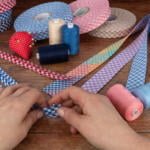 Hands working with checkered ribbons on a wooden table cluttered with various sewing supplies, including rolls of ribbons in different colors, spools of thread, scissors, and a pincushion shaped like a red strawberry.