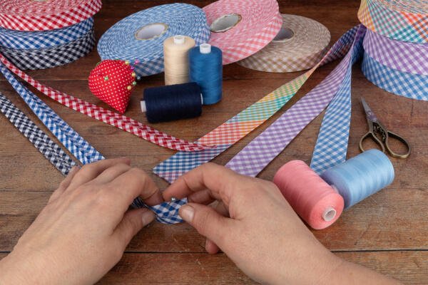 Hands working with checkered ribbons on a wooden table cluttered with various sewing supplies, including rolls of ribbons in different colors, spools of thread, scissors, and a pincushion shaped like a red strawberry.