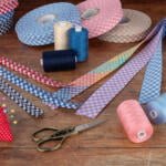 A wooden table is adorned with various sewing items, including multiple rolls of gingham ribbon, spools of thread in assorted colors, a red pincushion shaped like a strawberry, and a pair of vintage scissors. The setup suggests a sewing or crafting project.