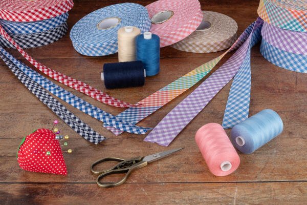 A wooden table is adorned with various sewing items, including multiple rolls of gingham ribbon, spools of thread in assorted colors, a red pincushion shaped like a strawberry, and a pair of vintage scissors. The setup suggests a sewing or crafting project.