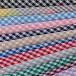 A stack of fabric rolls with a checkered pattern in various colors is arranged. The colors include black, yellow, turquoise, pink, blue, green, navy, beige, red, gray, and green. Each fabric has a white checkered pattern.