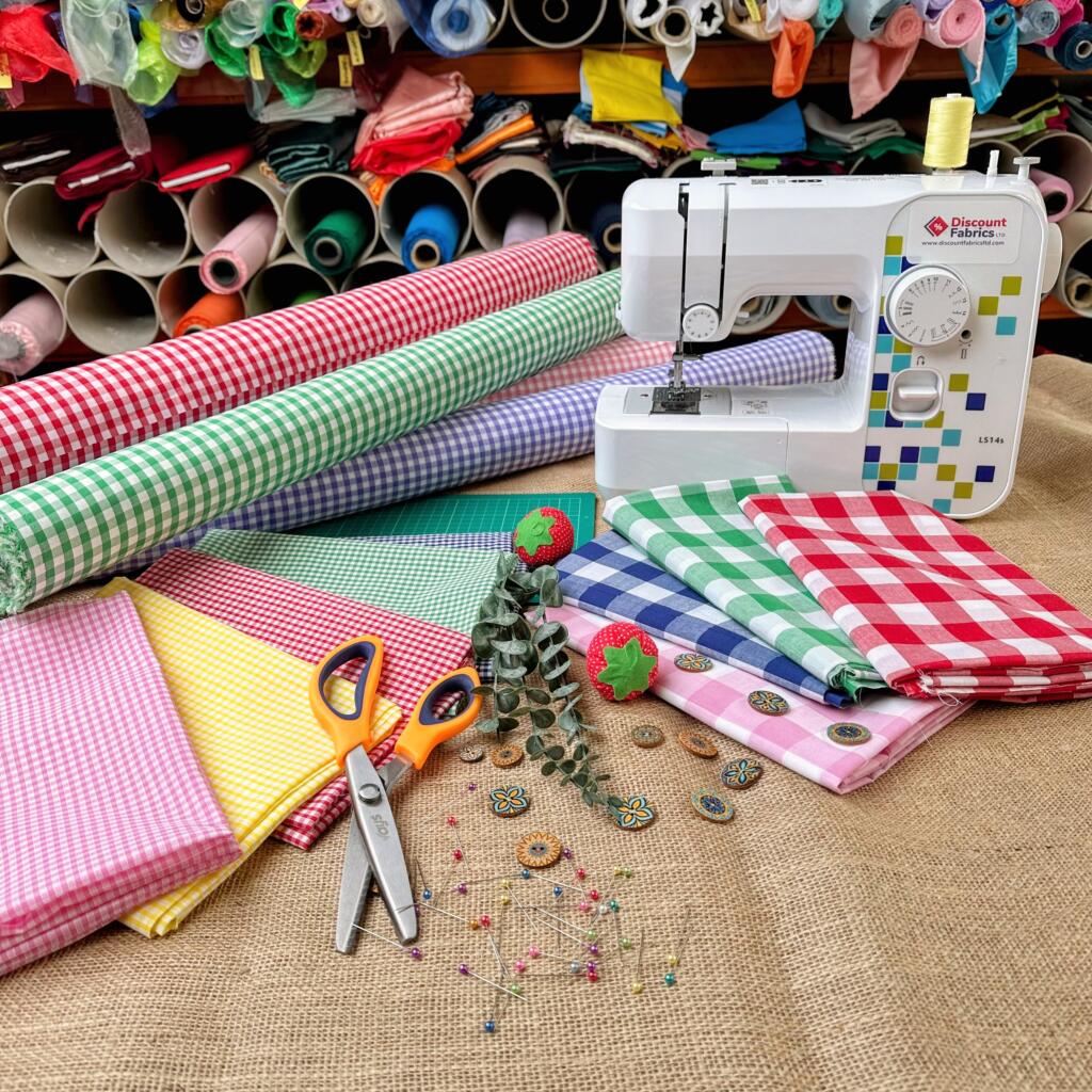 A sewing setup showing a white sewing machine surrounded by colorful rolls of fabric and folded fabric pieces in checkered patterns. Scissors, green and red pincushions, scattered buttons, pins, and threads are also displayed on a burlap surface. Rolls of fabric are stacked in the background.