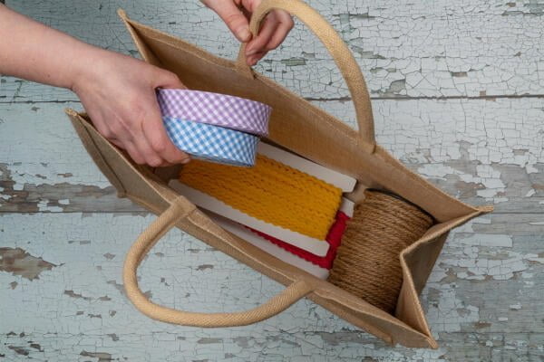 A person places two rolls of gingham ribbons, one blue and one purple, into a jute tote bag. The bag already contains several other crafting materials, including a bundle of yellow rope, a red item, and a spool of brown twine. The background is a distressed, painted wooden surface.