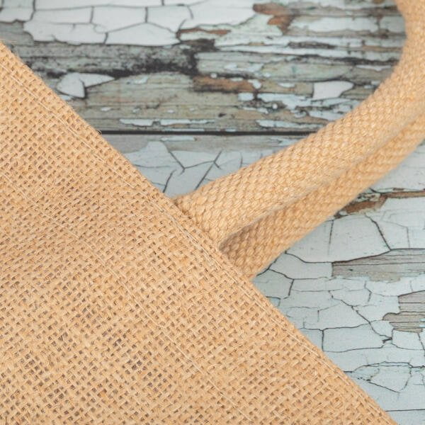 Close-up of a burlap tote bag handle resting on a distressed wood surface. The texture of the burlap and the peeling paint on the wooden background are clearly visible, adding a rustic appearance to the image.