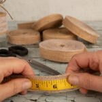 Two hands hold a yellow measuring tape against a strip of burlap ribbon with lace. Scissors rest beside a mason jar and various rolls of burlap ribbon in the background on a light-colored, textured table.