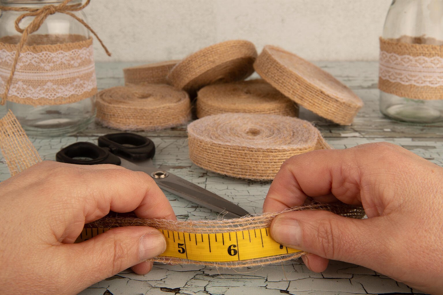 Two hands hold a yellow measuring tape against a strip of burlap ribbon with lace. Scissors rest beside a mason jar and various rolls of burlap ribbon in the background on a light-colored, textured table.