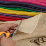 A person is cutting a piece of beige fabric using orange-handled scissors. Colorful fabric rolls in red, green, purple, and yellow are stacked in the background. Metal clips and rings are placed near the fabric being cut.