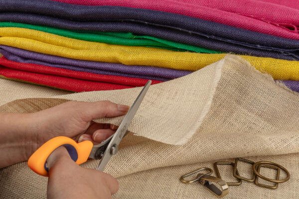 A person is cutting a piece of beige fabric using orange-handled scissors. Colorful fabric rolls in red, green, purple, and yellow are stacked in the background. Metal clips and rings are placed near the fabric being cut.