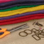 A stack of colorful fabric in purple, red, green, and yellow lies on a burlap surface. Nearby are a pair of orange-handled pinking shears, metal rings, rectangular clasps, small metal clips, and a few colorful pins.