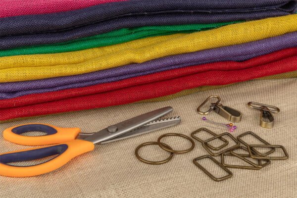 A stack of colorful fabric in purple, red, green, and yellow lies on a burlap surface. Nearby are a pair of orange-handled pinking shears, metal rings, rectangular clasps, small metal clips, and a few colorful pins.