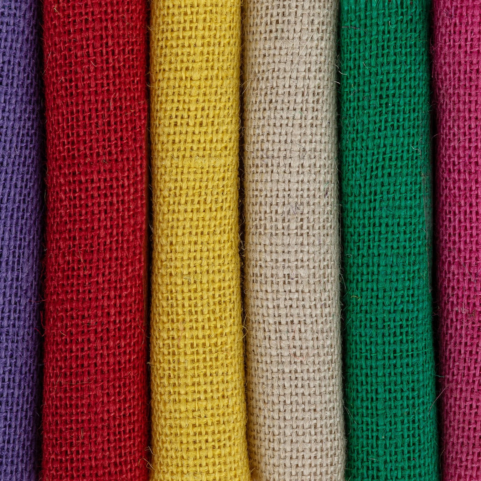 A close-up image shows six rolls of burlap fabric in different colors standing vertically. From left to right, the colors are purple, red, yellow, beige, green, and pink. The texture of the coarse weave is clearly visible in each roll.