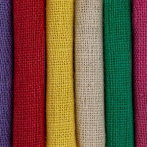 A close-up image shows six rolls of burlap fabric in different colors standing vertically. From left to right, the colors are purple, red, yellow, beige, green, and pink. The texture of the coarse weave is clearly visible in each roll.