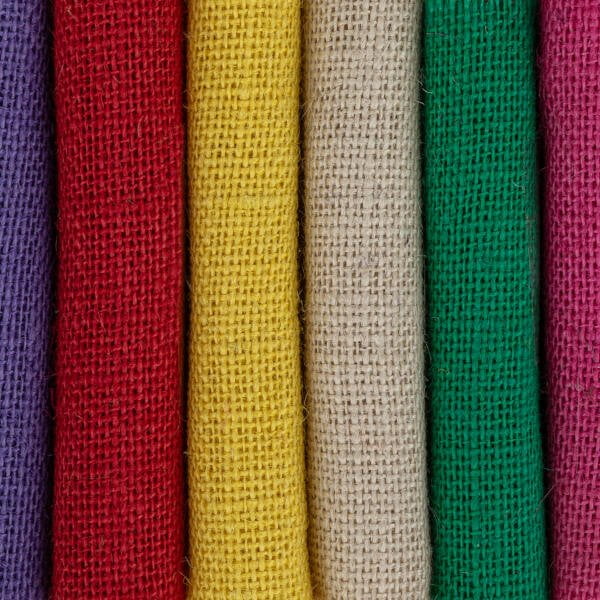 A close-up image shows six rolls of burlap fabric in different colors standing vertically. From left to right, the colors are purple, red, yellow, beige, green, and pink. The texture of the coarse weave is clearly visible in each roll.