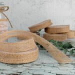 A rustic still life featuring glass jars and bottles adorned with burlap and lace ribbons. Coiled burlap ribbon is in the foreground on a weathered, cracked paint surface, with additional jars and ribbon spools arranged artistically in the background.