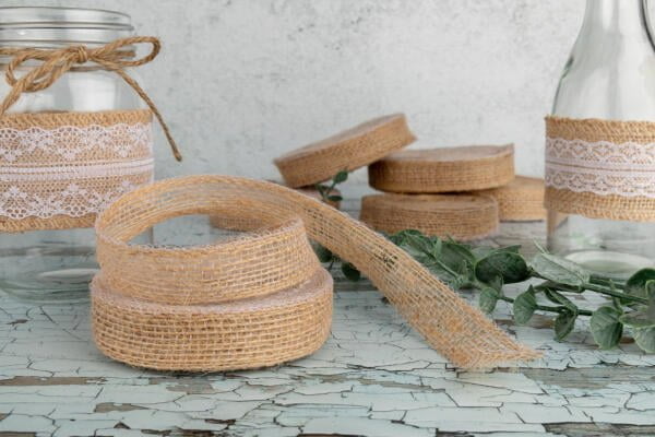 A rustic still life featuring glass jars and bottles adorned with burlap and lace ribbons. Coiled burlap ribbon is in the foreground on a weathered, cracked paint surface, with additional jars and ribbon spools arranged artistically in the background.