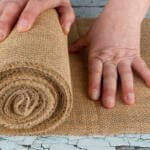 A person with light skin rolls out a piece of burlap fabric on a worn, chipped blue surface. The person's right hand holds the end of the rolled fabric while the left hand smooths it out, preparing it for use.