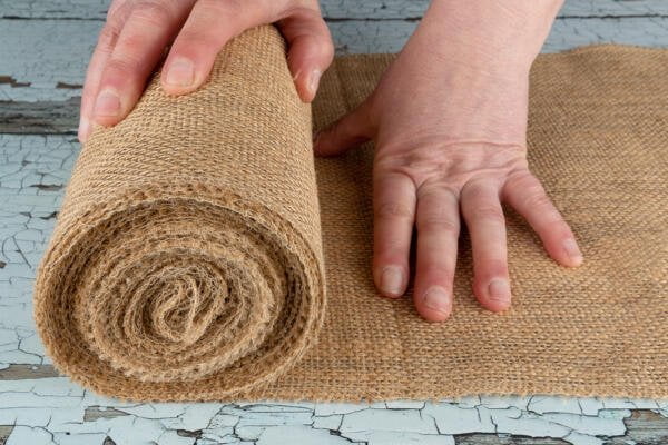 A person with light skin rolls out a piece of burlap fabric on a worn, chipped blue surface. The person's right hand holds the end of the rolled fabric while the left hand smooths it out, preparing it for use.