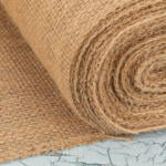 Close-up of a textured roll of burlap fabric, partially unrolled on a cracked surface. The fabric is a natural, coarse material with a light brown hue, prominently showing its woven pattern. The background has a weathered, pale blue appearance.