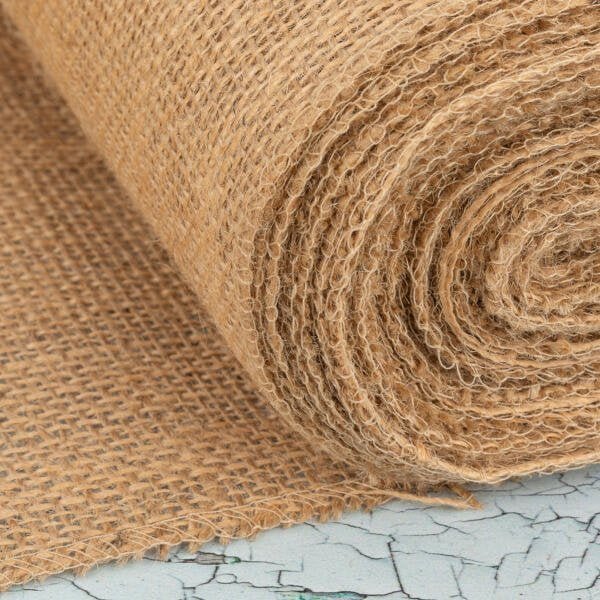 Close-up of a textured roll of burlap fabric, partially unrolled on a cracked surface. The fabric is a natural, coarse material with a light brown hue, prominently showing its woven pattern. The background has a weathered, pale blue appearance.