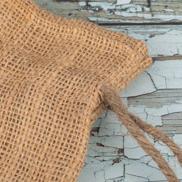 Close-up of a textured burlap drawstring bag on a weathered, cracked wooden surface. The drawstring is partially pulled out, emphasizing the rustic and natural materials of the bag.
