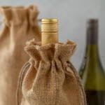 Two bottles of wine, one with a golden cap and the other mostly hidden, are each wrapped in burlap sacks with drawstrings. The focus is on the golden-capped bottle in the foreground, while the background bottle is blurred. The setting is minimalist.