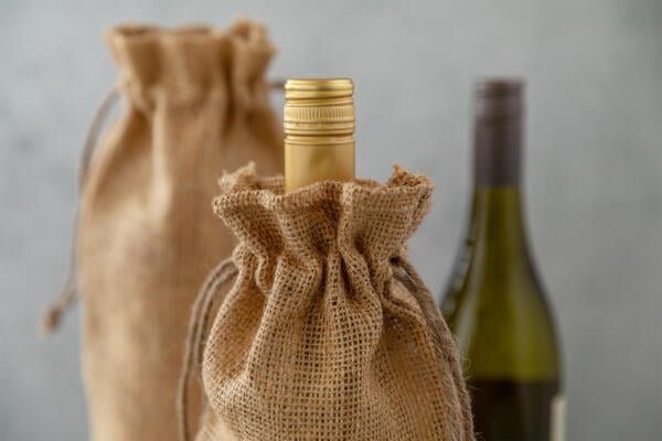 Two bottles of wine, one with a golden cap and the other mostly hidden, are each wrapped in burlap sacks with drawstrings. The focus is on the golden-capped bottle in the foreground, while the background bottle is blurred. The setting is minimalist.