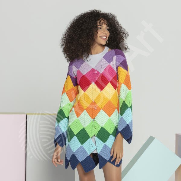 A person with curly hair smiles while modeling a colorful, diamond-patterned, knitted cardigan that features various colors including purple, red, yellow, green, and blue. The background is minimalistic with pastel-colored blocks.