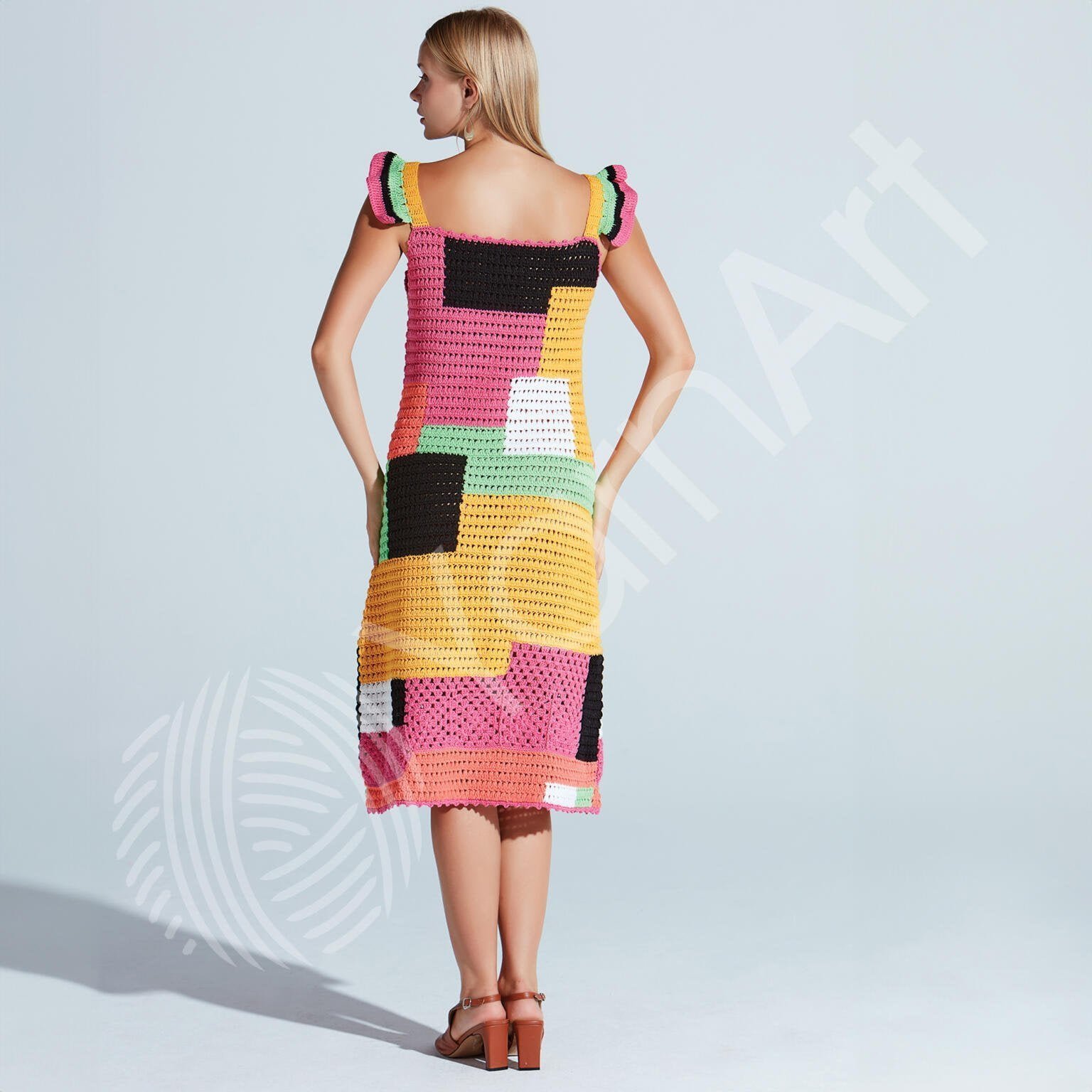 A woman with blonde hair is standing with her back to the camera, wearing a colorful patchwork crochet dress with block patterns in orange, pink, yellow, green, black, and white. She is also wearing brown sandals and stands against a light grey background.