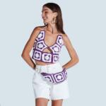 A woman stands confidently against a plain background, wearing a purple and white crocheted halter top and matching belt with a pouch. She is also wearing high-waisted white shorts. Her hair is down and she smiles while looking off to the side.