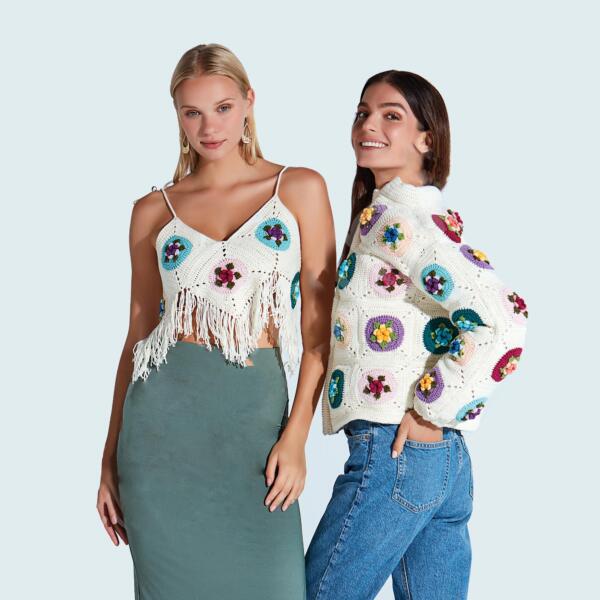 Two women pose against a light background. One on the left wears a white crocheted top with a fringe and a long, green skirt. The other on the right wears a white crocheted jacket with colorful circular patterns, paired with blue jeans. Both are smiling.
