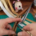Close-up of hands cutting gold glitter ribbon with scissors on a green cutting mat. Surrounding the hands are various rolls of metallic ribbon, gray thread bobbins, and a fabric pincushion shaped like an animal with pins on its back.